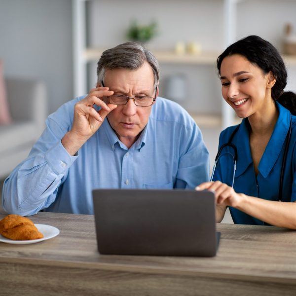 Millennial caregiver showing elderly disabled man in wheelchair how to use laptop at retirement home. Young female doctor helping senior male patient to find information on web indoors